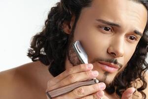 Man with curly hair grooming his beard carefully using a straight razor against a plain white background photo