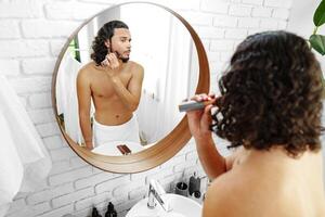 A man with curly hair grooming himself in the bathroom while looking into a round mirror photo