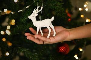 New Year and Christmas greeting card with a deer. A hand holds a deer toy near the Christmas tree. photo