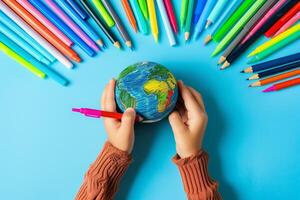 Child's hands holding a colorful globe surrounded by pencils and markers on a blue background photo