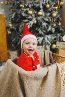 A little boy 1 year old sits in a basket against the background of a Christmas tree. A child in a red New Year's wind near the Christmas tree photo