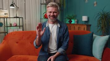 Mature man smiling friendly at camera, waving hands gesturing hello, hi, greeting at home on couch video
