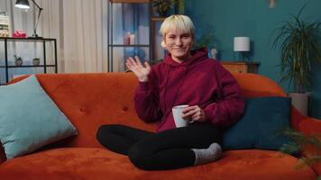 Young woman smiling friendly at camera, waving hands gesturing hello, hi, greeting at home on couch video