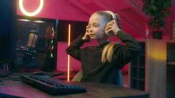Teenager little girl gamer streamer put on headphones looking at camera hand greeting hello hi prepare start streaming daughter child kid playing computer play virtual game online at home neon video