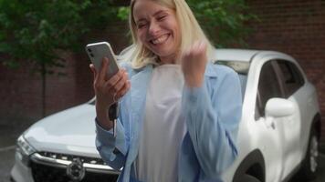 Joyful surprised Caucasian woman buyer female smile using mobile phone winning achievement car vehicle in city client businesswoman celebrating success girl automobile driver rejoicing buying new auto video
