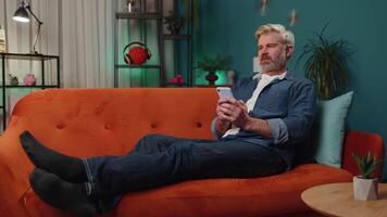 Portrait of mature man lying on sofa, using smartphone share messages on social media application video