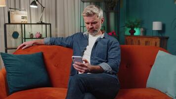 Middle-aged old man sitting on sofa, using smartphone share messages on social media application video
