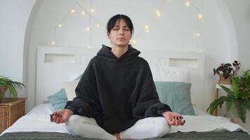 yoga consapevolezza meditazione. giovane salutare donna praticante yoga nel Camera da letto a casa. donna seduta nel loto posa su letto Meditare sorridente rilassante interno. ragazza fare respirazione la pratica. yoga a casa video