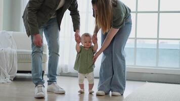 Baby development. Little infant girl making first steps at home. Happy mother father looking at toddler child walking in living room. Happy family at home. Mom dad supporting baby first steps video