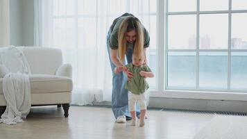 Baby development. First steps. Little infant girl making first steps at home. Happy mother supporting baby first steps. Mom helping toddler child walking in living room. Happy family at home video