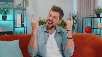 Overjoyed young man showing rock n roll gesture, cool sign dancing rejoicing in success at home video