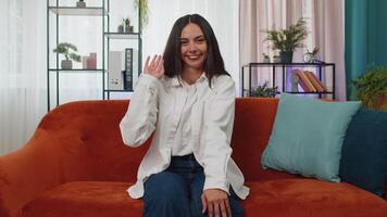 Young woman smiling friendly at camera, waving hands gesturing hello, hi, greeting at home on couch video