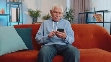 Senior old grandfather sitting on sofa, using smartphone share messages on social media application video