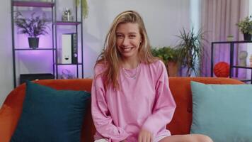 Young woman smiling friendly at camera, waving hands gesturing hello, hi, greeting at home on couch video