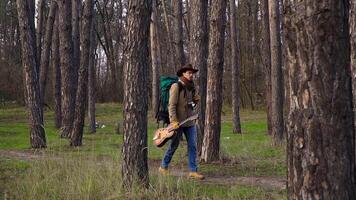 A backpacker with a guitar and a camera walks along a trail in the forest. Travel concept. Slow motion video