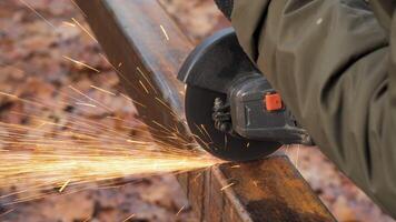 A worker uses an angle grinder to cut through a piece of metal, creating a shower of sparks against a backdrop of natural materials. The setting is rustic and practical. video