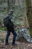 Man in camouflage clothing with survival backpack standing in front of self-built shelter in forest photo