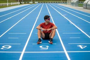 Resting Male Runner with Sunglasses on Ground at Finish Line of Blue Athletic Track After Long Race photo