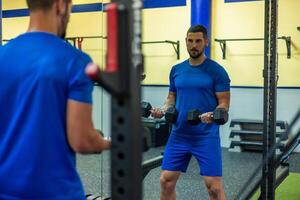 young fitness athlete doing bicep exercises with dumbbells while reflecting in gym mirror photo