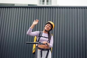 food delivery girl on electric scooter with yellow backpack and helmet greeting with raised hand photo