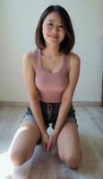 A smiling Asian woman with shoulder-length hair, wearing a pink tank top and denim shorts, sits on a floor. photo