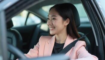 A smiling Asian woman in a pink blazer sits in the driver's seat of a car, holding the steering wheel. photo