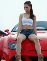 A young Asian woman with a slender physique, wearing a white crop top and denim shorts, sits on a red sports car. photo