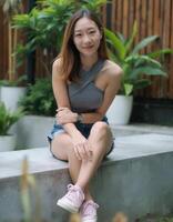 A smiling Asian woman with shoulder-length brown hair, wearing a grey halter top and denim shorts, sits on a bench. photo