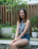 A smiling Asian woman with shoulder-length brown hair, wearing a grey halter top and denim shorts, sits on a bench. photo