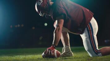 Portrait of an American football player at night at stadium lights with a helmet and protective gear uniform standing on one knee on the field holding the ball. Cinematic slow motion side view. video