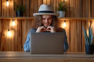 Man with long hair and hat forms a heart with hands during a call at a cozy wooden table photo