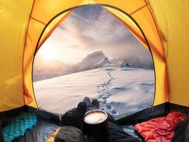 Man holding coffee cup and enjoying view of sunrise on snowy mountain inside yellow tent photo