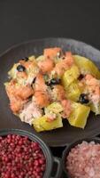 Baked salmon pieces with potatoes, cheese and olives with salt, spices and herbs on a dark concrete background video