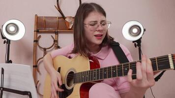 Young woman in eyeglasses plays six string acoustic guitar and sings song with so much emotion. She plucks strings like she's trying to break them, and shakes head to pulsating beat of music. Zoom in video