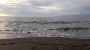 A serene beach view with gentle waves touching the shore under a cloudy sky, as the golden hues of a setting sun break through the horizon. video