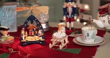 Person pouring tea into a cup on a table. Hands pour tea from teapot in cup on holiday table. Festively arranged decorations include nutcracker figures and miniature sled with marshmallows video