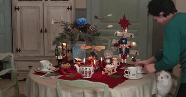 Woman put white cat on chair at holiday table. Warm home is adorned with festive decorations, featuring a beautifully set table with tea cups, figurines, and a playful cat exploring the surroundings. video