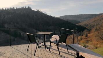 Scenic balcony with mountain views - A wooden balcony with a table and chairs overlooking a stunning valley surrounded by forested hills under a bright sky. video