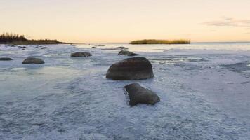A serene winter scene of a frozen shoreline along Lake Ladoga with scattered rocks partially covered by ice, reflecting the soft hues of sunset in the distance video