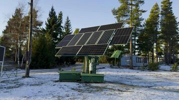 A large solar panel array stands in a snow-covered clearing surrounded by tall pine trees and wooden houses. The sunlight reflects off the panels, highlighting sustainable energy use even in winter video