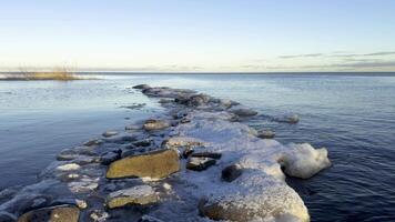 A serene winter view of the frozen shoreline of Lake Ladoga at sunset. Snow and ice cover the rocky shore, while the calm water reflects the golden light of the setting sun, creating a peaceful video