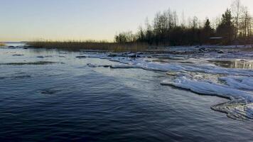 A serene winter view of the frozen shoreline of Lake Ladoga at sunset. Snow and ice cover the rocky shore, while the calm water reflects the golden light of the setting sun, creating a peaceful video