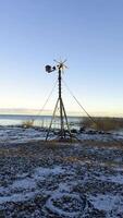 A close-up of a weathered windmill standing on the snowy shore of Lake Ladoga, with calm water and reeds in the background. The sunset light highlights the rustic structure against the winter video