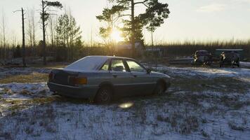 An old car sits idle and dusted with snow in a rural winter setting, with the golden glow of the sunset casting long shadows. Boats and wooden structures can be seen in the background, adding to the video