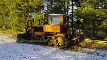An aged orange bulldozer with rusted parts stands idle in a snow-covered forest clearing. The winter sunlight highlights the machinery, surrounded by tall pine trees, evoking a sense of abandonment video