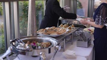 A formal event featuring a buffet with diverse food items, including flatbreads and falafel wraps. A person in gloves serves food to a formally dressed attendee. Ideal for event and catering video