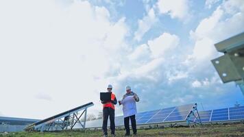 Engineers discussing blueprint on a laptop. Successful workers in protective uniform looking at plan on the background of solar panels farm. video