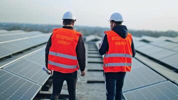 Alternative green energy on the solar station. Backside view of two workers on the electricity farm. Technicians in protective uniform and helmets. video
