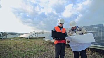 Technicians look into laptop and project plan on the solar farm. Two workers in protective helmets on the background of solar panels. video