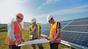Installation of solar panels. Three workers looking at the plan on a solar farm in summer. Blue solar batteries on the field for renewable energy. video